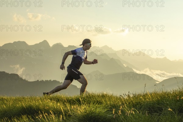 Man in athletic gear running on a grass trail with scenic mountain backdrop. The sun sets behind the peaks, casting a warm glow. Wearing a hat and sunglasses, he embodies outdoor activity