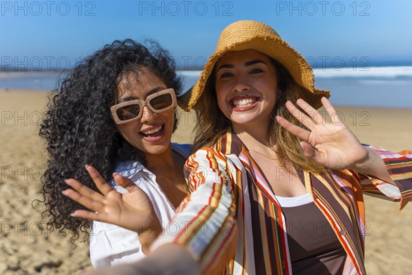 Two cheerful young women on a sunny beach vacation take a joyful selfie, smiling and waving at the camera with sunhats and sunglasses, relaxed and carefree together