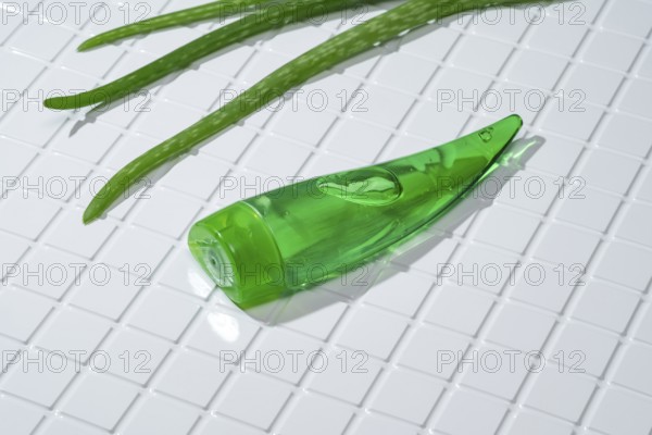 A green aloe vera gel bottle lies alongside fresh aloe leaves on a clean, white-tiled bathroom surface, depicting natural skincare products