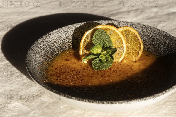 A caramel dessert elegantly served in a dark textured bowl, topped with dried orange slices and fresh mint leaves, casting a soft shadow under warm lighting