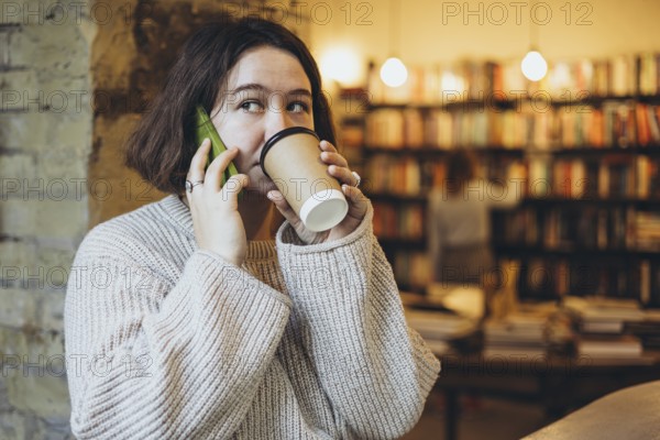 A young woman in a cozy sweater drinks coffee while talking on her smartphone inside a warm bookstore cafe The shelves filled with books create a relaxed and inviting atmosphere