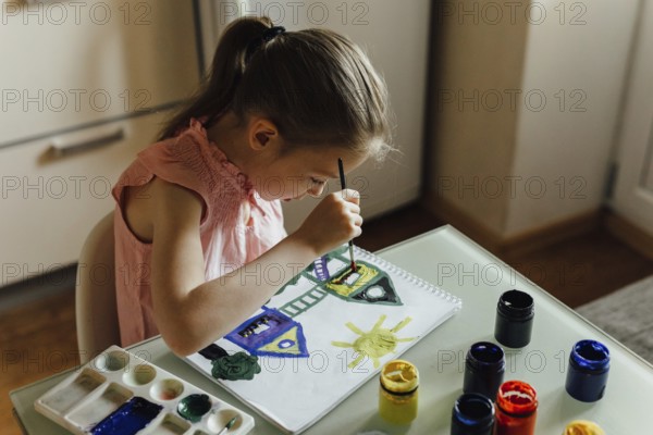 A young girl wearing a pink outfit sits at a clear table, focused intently as she paints a colorful house and sun with brushes and jars of vibrant paint surrounding her