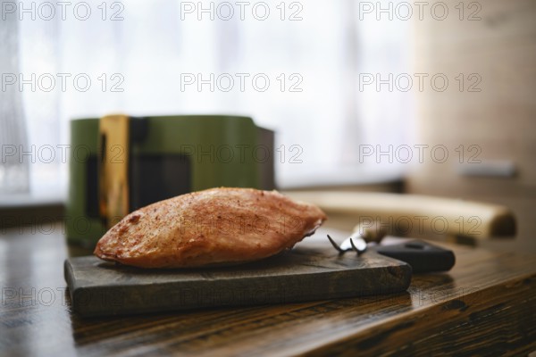 Roasted pork fillet rests on a wooden cutting board in a kitchen table. Air fryer basket and kitchen tools are on blurred background. Natural light fills the room enhancing the cooking scene