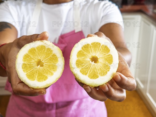 A woman in a pink apron presents two fresh lemon halves in a kitchen setting. The vivid yellow citrus contrasts with the neutral background, enhancing the fresh, vibrant feel