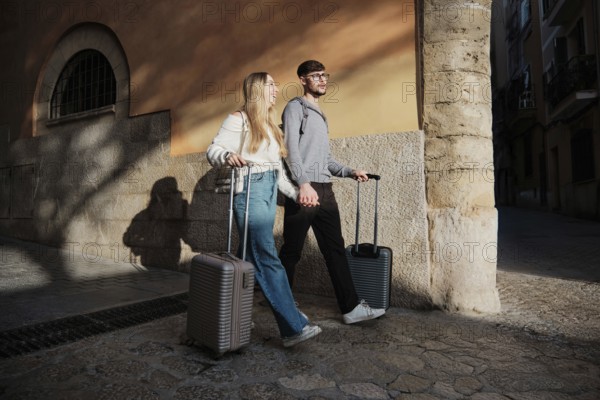 A young couple strolls through a picturesque street in Majorca, Spain. They hold hands and pull suitcases, excitedly exploring potential vacation homes