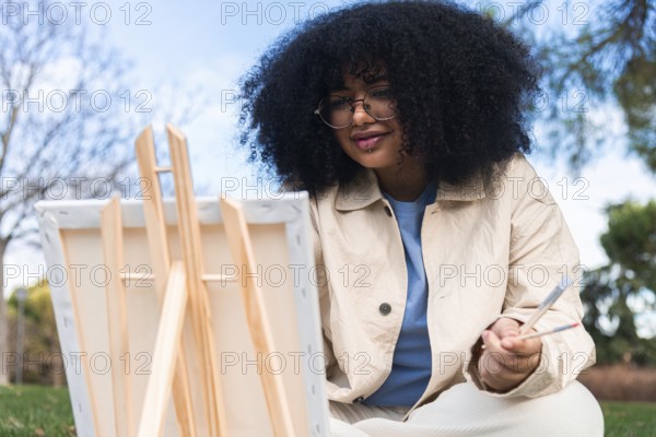 A young black woman enjoys painting outdoors in a sunny park, capturing the beauty of nature. She holds brushes and focuses on her canvas, surrounded by vibrant scenery
