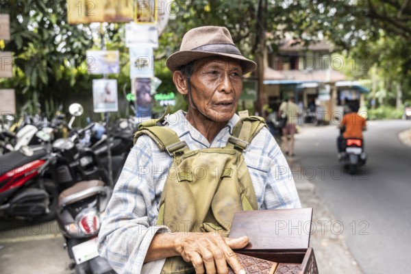 An elderly balinese man wearing a hat and plaid shirt stands on a street, holding a wooden box. Scooters and greenery surround him, creating a vibrant urban scene