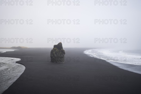A haunting view of Reynisfjara Beach, Iceland, featuring a solitary rock formation amidst the black volcanic sand under a misty winter sky, captured in solitude