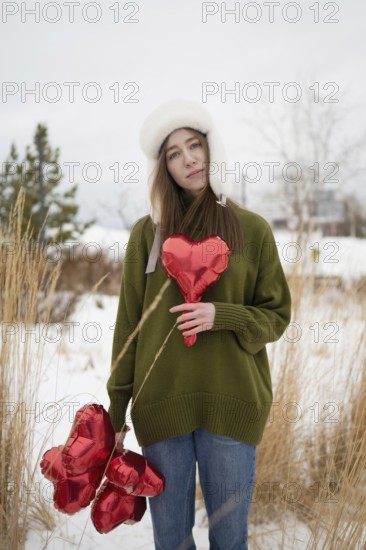 Woman in a green sweater, holding red heart shaped balloons in a snowy park, surrounded by dry grass, embodying warmth and love against cold