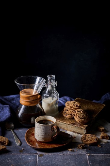 A cozy setup featuring a rustic ceramic cup of coffee, a glass bottle of milk, and a stack of delicious chocolate and butter cookies on an old book, evoking warm, inviting vibes
