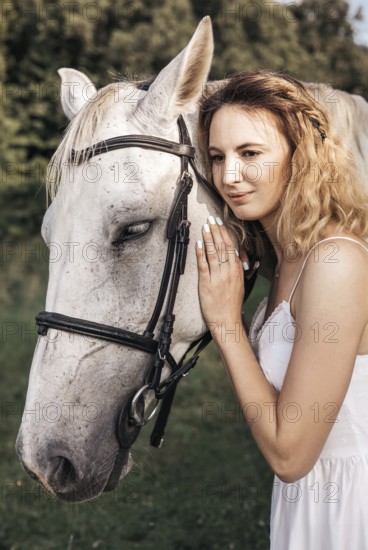 A young woman in a white dress gently embraces a serene white horse outdoors, showcasing a peaceful connection between human and animal on a sunny day