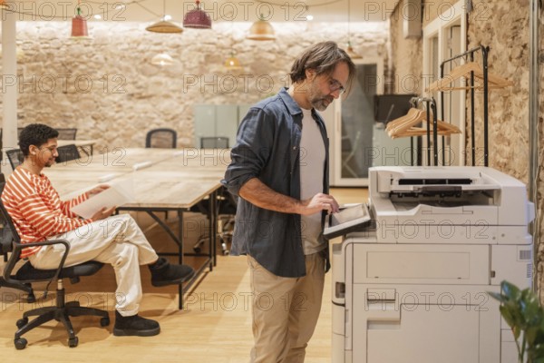 Two professionals in a cozy coworking space, one seated with documents and the other using a copier. The rustic setting exudes creativity and collaboration