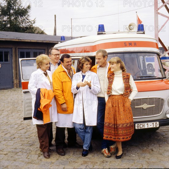 Die Schauspieler v.l.: Ingeborg Krabbe, Otmar Richter, Günter Schubert, Uta Schorn, Wilfried Pucher und Anne Kasprik vor einem DMH-Krankenwagen in einer Szene der DDR-Fernsehserie "Bereitschaft Dr. Federau", undatiertes Foto von 1988. Die siebenteilige Serie um Dr. Uta Federau (Uta Schonr) war eine der wenigen in den sozialistischen Ländern produzierten Arztserien.