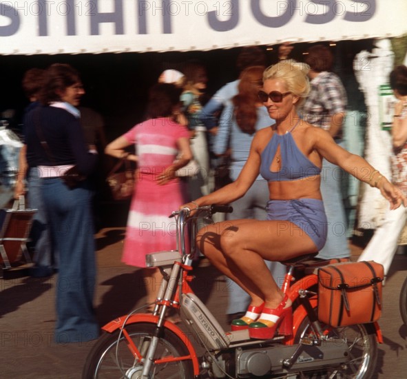 Where the rich and beautiful go on holiday: A woman, lightly dressed for summer and wearing high-fashion shoes with platform soles, rides a moped through St Tropez in France. Taken in 1975.