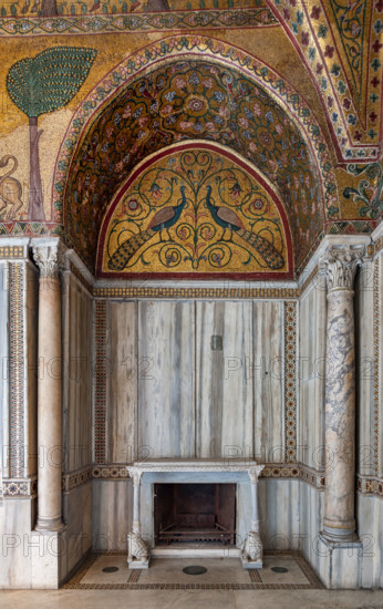Palermo, Palazzo Reale o Palazzo dei Normanni, Torre Pisana, Sala di Re Ruggero: veduta della sala voluta da Re Ruggero II d'Altavilla. E' caratterizzata da ampie superfici a mosaico di grande pregio raffiguranti elementi vegetali (palme e banani) e scene di carattere aulico e venatorio, simboli del potere normanno (battute di caccia con arcieri e cervi, rappresentati pavoni, cigni, oltre i mitologici centauri, grifi e altri animali esotici tra cui leopardi e tigri fra lussureggiante vegetazione). I mosaici sono di stile greco - bizantino (secolo XIII).
Particolare di nicchia con caminetto con due leoni stilofori che reggono la mensola, lunetta con mosaico di pavoni; ricchi rivestimenti in marmo e colonnine angolari.

[ENG]
Palermo, The Royal Palace or Palazzo dei Normanni (Palace of the Normans), Pisana Tower,  the King Roger Room: view of the Room which was commissioned by King Roger II d'Altavilla. It is characterized by large mosaic surfaces of great value representing plant el...