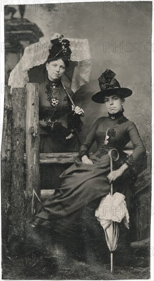 Portrait of Two Adult Women, One Seated, One Standing, in Hats and Long Dresses and Holding Parasols, circa 1870