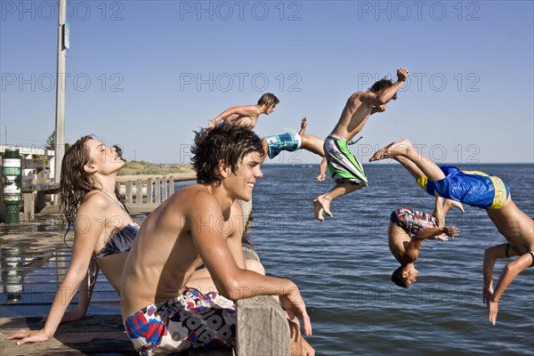 Young Couple Sitting on Edge of Pier With Group of Boys Jumping in Water