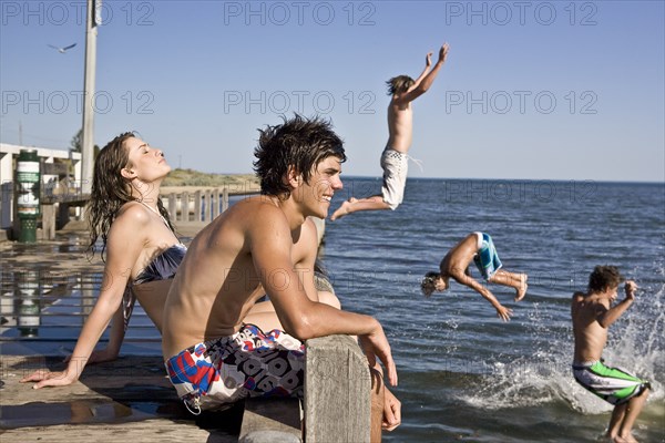 Young Couple Sitting on Edge of Pier With Group of Boys Jumping in Water