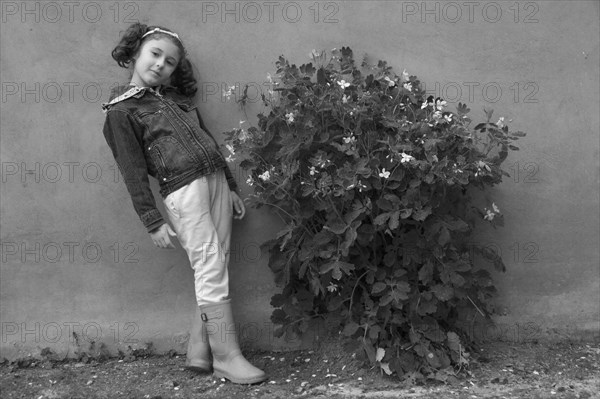 Young Girl Standing Against Wall Next to Flowering Plant