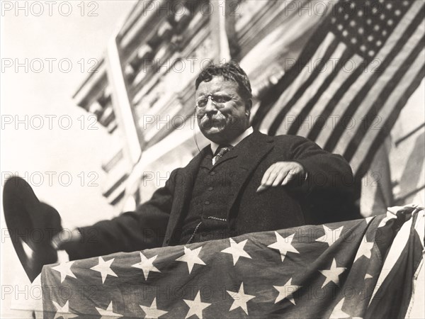 Theodore Roosevelt on a flag-draped platform, with hat in hand during his tour through New Jersey before the Progressive Party Convention, September 1912