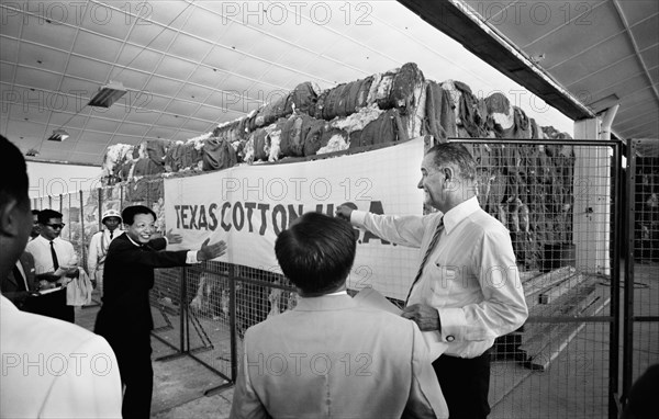 U.S. Vice President Lyndon Johnson Visiting Textile Mill with Vietnamese men standing in front of a large quantity of textiles with a "Texas Cotton U.S.A." Banner, Saigon, South Vietnam, photograph by Thomas J. O'Halloran, May 1961