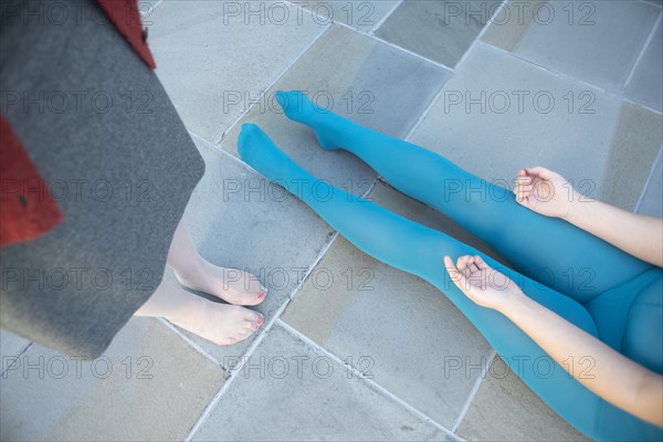 High Angle View of Two Women, One in Blue Unitard Sitting on Ground with Legs Extended, while the other Stands Nearby in Dress and Mesh Stockings, View from Waist-Down