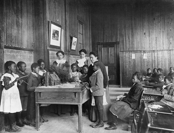 African American Children learning about Thanksgiving, with model Log Cabin on Table, 1900