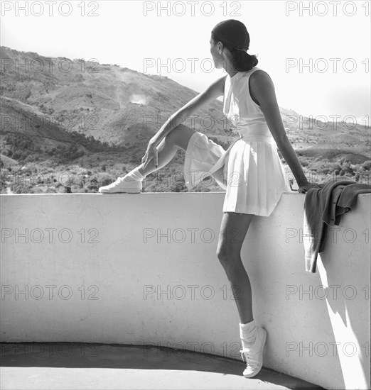 Fashion Model wearing Tennis Outfit, Full-Length Portrait sitting on Wall with Mountains in Background