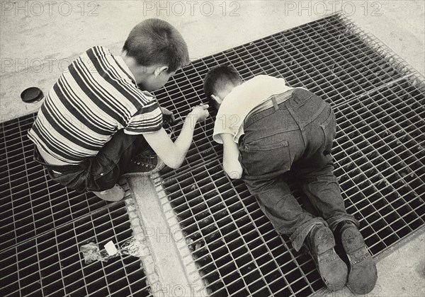 Two Young Boys peering through Subway Grate on Sidewalk while lowering a String with a fastener to snag a Prize