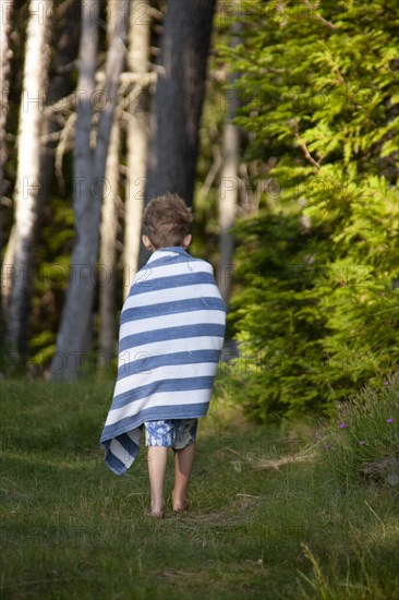 Rear view of young boy walking along grassy path wrapped in striped towel