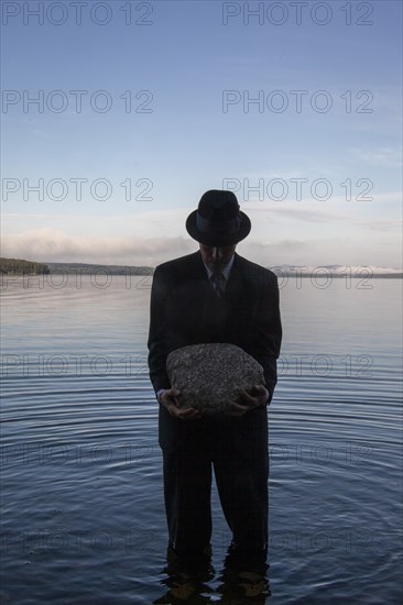 Man in suit and hat holding boulder while standing in calm lake at sunset