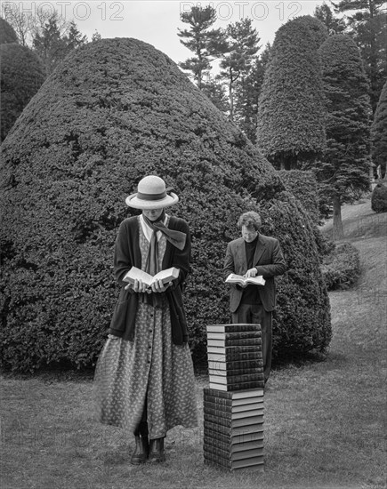 Man and woman reading while standing near stack of books in topiary garden