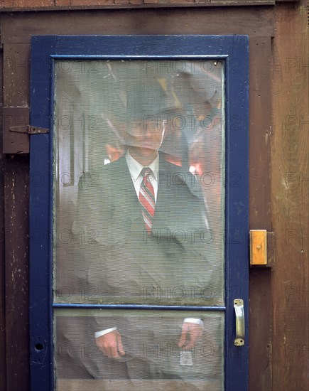 Man in suit and hat standing behind rustic screen door