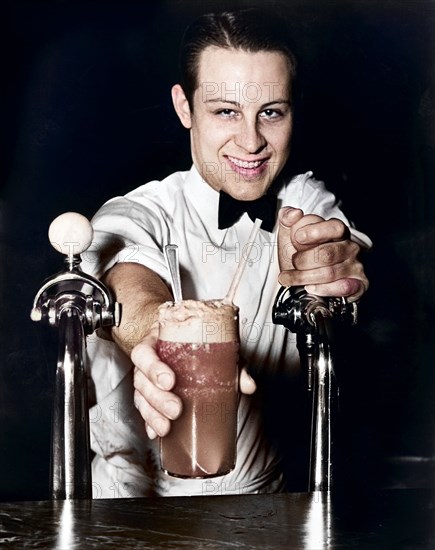 Soda jerk passing ice cream soda between two soda fountains