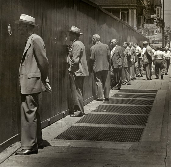 Row of men standing on sidewalk looking through round peep holes at new construction site