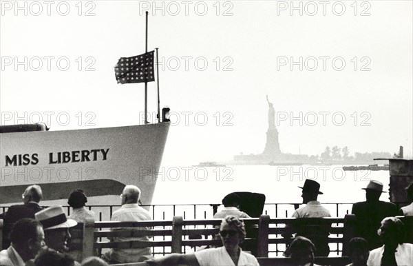Group of people sitting on benches facing Statue of Liberty with boat named 'Miss Liberty' is on  left
