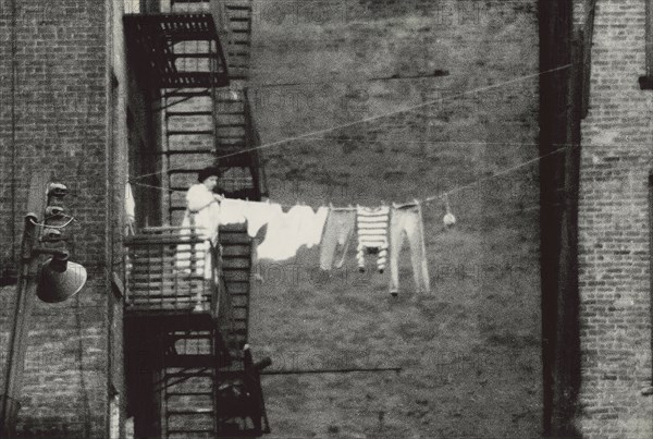 Woman standing on fire escape hanging clothes to dry on clothes line running to neighboring building