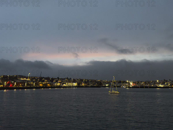 Sailboat with Christmas lights in harbor with illuminated seaside town in background, Hafnarfjordur, Iceland