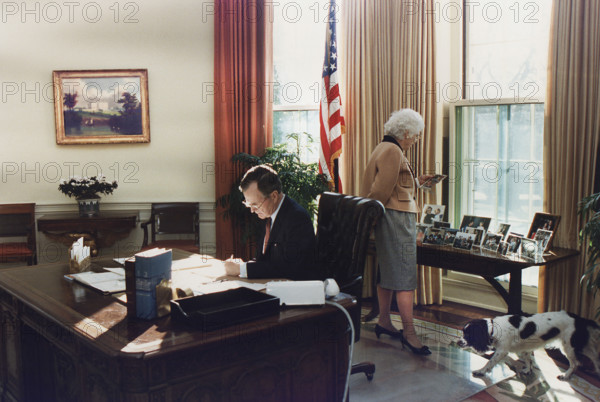 U.S. President George H.W. Bush working at his desk in the Oval Office as U.S. First Lady Barbara Bush looks at photographs on the table behind desk, White House, Washington, D.C., USA, Susan Biddle, White House photographer, January 27, 1989