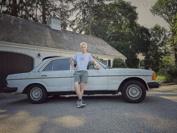 Young adult man with bleached hair leaning against vintage Mercedes car in driveway