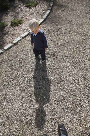 High angle view of young boy casting long shadow on gravel driveway