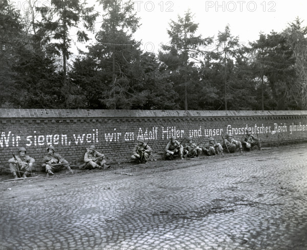 Members of 16th regiment, 1st Division, U.S. First Army, sitting under German slogan "Wir Siegen, weil wir an Adolf Hitler und unser Grossdeutches Reich Glauben", English translation, "We triumph because we believe in Adolf Hitler and our Greater German Reich", Vetteweiss, Germany, U.S, Army Signal Corps, February 28, 1945