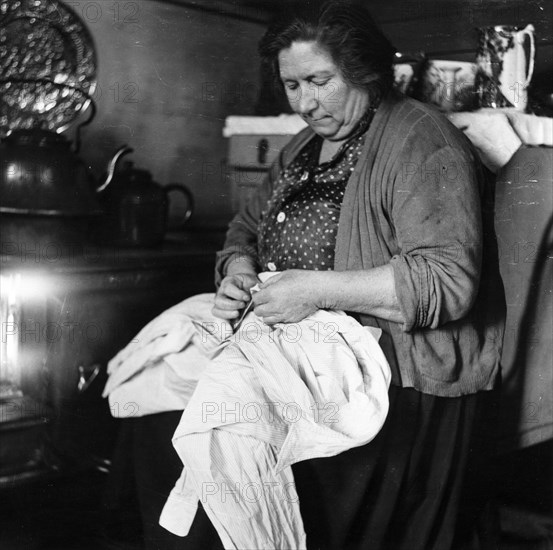 A woman sewing a shirt by the stove, London, 1953. Artist: Henry Grant