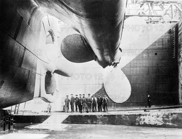 Workmen standing under one of the propellors of the Titanic, 31 May, 1911 (b/w photo)