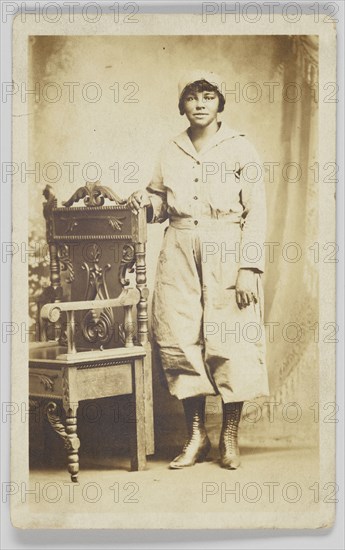 Photographic postcard of a woman standing next to a wooden chair, 1918-1930. Creator: Unknown.