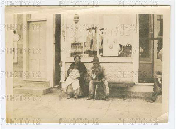 Photograph of a man and woman sitting outside of a storefront, early 20th century. Creator: Unknown.