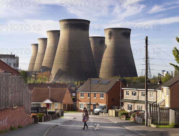 Ferrybridge C Power Station, West Yorkshire, 2018. General view looking north-west along a suburban street, with a woman walking a dog in the foreground and the power station's cooling towers dominating the skyline behind.
