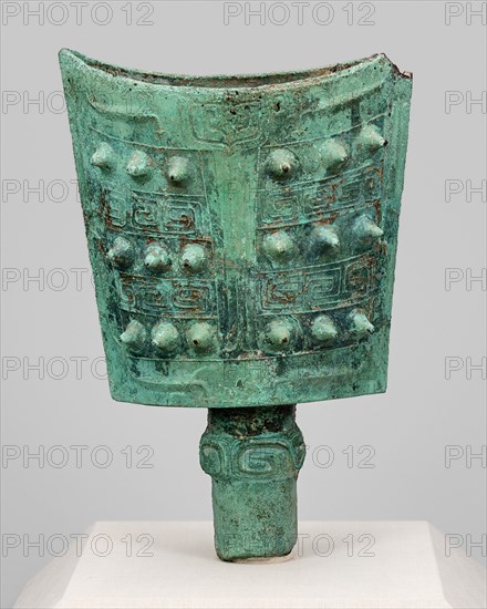 Bell (nao), Western Zhou dynasty (1046-771 B.C.). A bell with two convex sides of nearly rectangular shape and an opening at top rests on a slim cylindrical base. It boasts a green patina and is decorated with a geometric pattern and spikes. A small portion is missing at upper right.