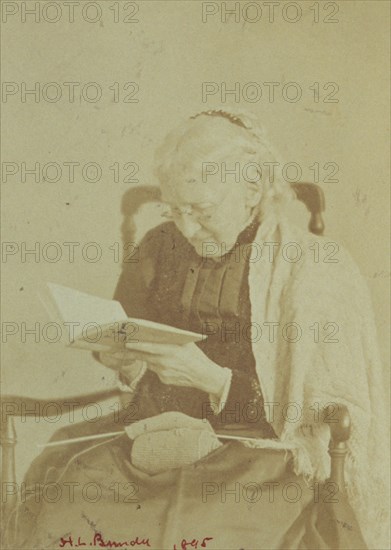 Elderly woman seated and reading a book with knitting in her lap, 1895.
