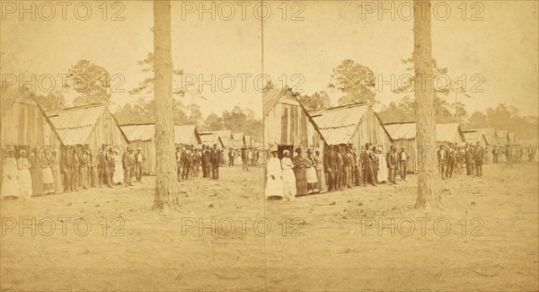 Group lined up in front of a row of shacks, (1868-1900?). Creator: Unknown.